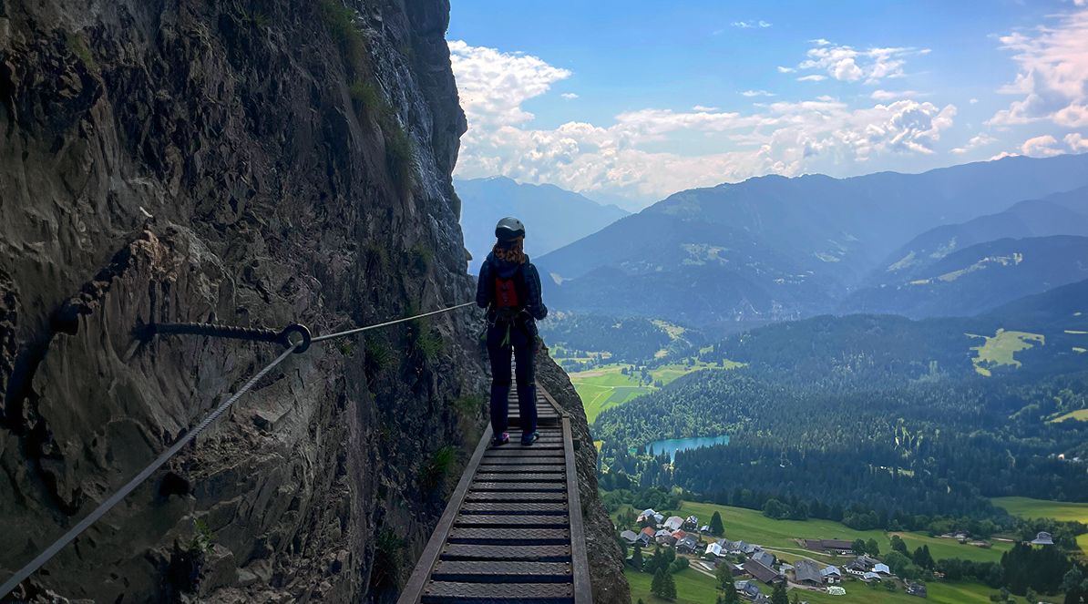 Eine Bergsteigerin in Kletterausrüstung steht auf einem schmalen Klettersteig an einer Felswand links im Bild, die nach rechts unmittelbar neben der Bergsteigerin vertikal abfällt. Im Vordergrund ist die Sicherungsvorrichtung in der Felswand zu sehen. Die