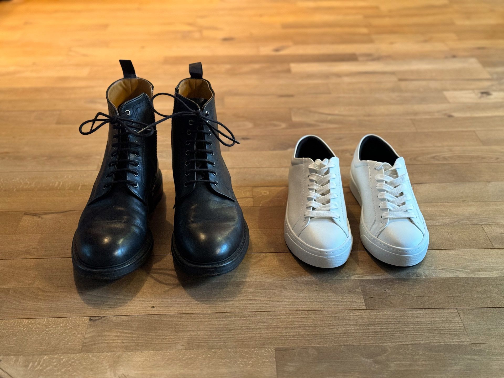 Ein Paar schwarze Herrenschnürstiefel steht neben einem Paar weißen Damen-Leder-Sneakern auf einem Holzfußboden.